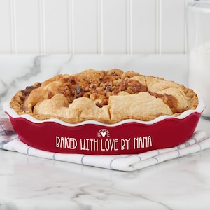 May include: Red ceramic pie dish with a white rim and the text "Baked with love by Nana" in white. The dish is filled with a baked apple pie with a golden brown crust and a topping of chopped walnuts.