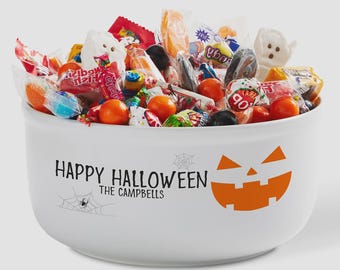 May include: A white ceramic bowl filled with assorted colorful candies. The bowl features an orange jack-o'-lantern face, a spider web graphic, and the black printed text "HAPPY HALLOWEEN THE CAMPBELLS" on the front exterior surface.