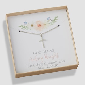 May include: A silver cross pendant on a delicate chain inside a square cardboard gift box. The insert features a floral illustration and the text: GOD BLESS Audrey Knight First Holy Communion May 12, 2026, printed in gray and pink script.