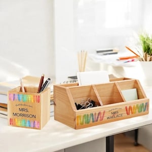 May include: Two wooden desk organizers with rainbow crayon designs. The smaller organizer is a pencil holder with the text "Best teacher ever MRS. MORRISON". The larger organizer has three compartments and the text "Best teacher ever MRS. WALLACE".