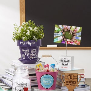 May include: A collection of teacher appreciation gifts including a purple flower pot with the text "Mrs. Flynn" and "thank you for helping me grow", a pink bucket with the text "Miss Tina You Are One Sweet Teacher", a wooden trophy with the text "1st Teacher Mr. Harper", a glass jar with the text "Teaching is a work of heart! Mrs. Miller Lisle Junior High School", and a white sign with the text "It takes a big heart to handle little minds Mrs. Enriquez".