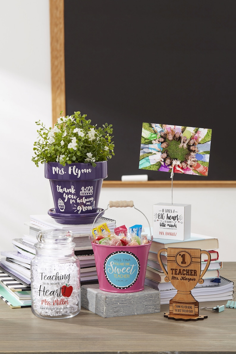 May include: A collection of teacher appreciation gifts including a purple flower pot with the text "Mrs. Flynn" and "thank you for helping me grow", a pink bucket with the text "Miss Tina You Are One Sweet Teacher", a wooden trophy with the text "1st Teacher Mr. Harper", a glass jar with the text "Teaching is a work of heart! Mrs. Miller Lisle Junior High School", and a white sign with the text "It takes a big heart to handle little minds Mrs. Enriquez".