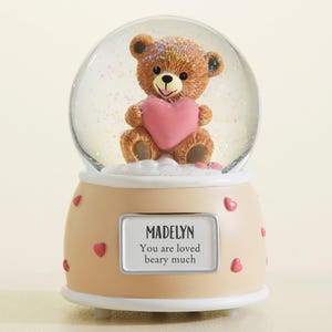 May include: A snow globe featuring a brown teddy bear holding a pink heart. The bear is inside a clear glass globe filled with glitter. The base is tan with pink hearts and a label that reads "MADELYN You are loved beary much."