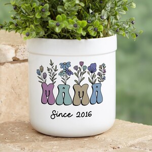 May include: White ceramic planter with a floral design and the word "MAMA" in colorful letters. The planter also has the text "Since 2016" below the word "MAMA".