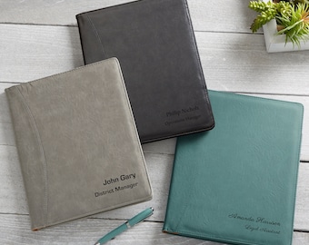 May include: Three personalized leather portfolios in gray, charcoal, and teal, each with a name and title. A teal pen rests near the portfolios. The portfolios are on a light wood surface.