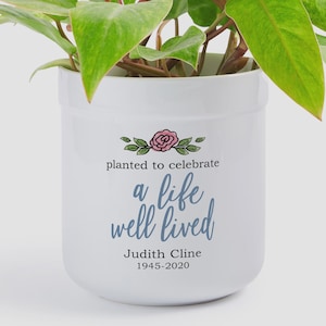May include: A white ceramic cylindrical planter containing a green leafy plant. The front features a pink rose illustration above the text "planted to celebrate a life well lived" and the name "Judith Cline" with the years "1945-2020" below.