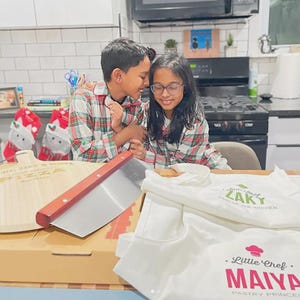 May include: A wooden pizza peel with the text Zaky & Maiya Pizza, a metal pizza cutter with a wooden handle, and two white aprons. One apron reads Little Chef Zaky King of the Kitchen, and the other reads Little Chef Maiya Pastry Princess.