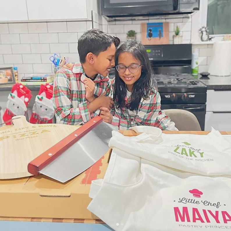 May include: A wooden pizza peel with the text Zaky & Maiya Pizza, a metal pizza cutter with a wooden handle, and two white aprons. One apron reads Little Chef Zaky King of the Kitchen, and the other reads Little Chef Maiya Pastry Princess.