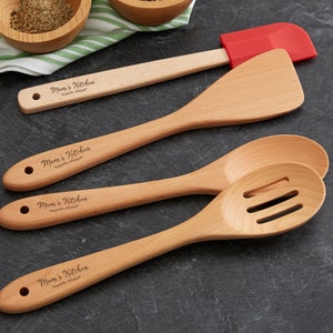 May include: Set of four wooden kitchen utensils with the text "Mom's Kitchen" engraved on each. The set includes a spatula, a slotted spoon, a solid spoon, and a red silicone spatula.