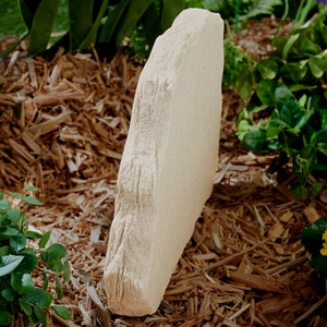 May include: A beige, rectangular garden stone with a rough, textured surface. The stone is standing upright in a bed of wood chips, surrounded by green plants. The stone appears to be made of sandstone.