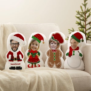 May include: Four white Christmas-themed pillows with faces of people in the center. The pillows are shaped like Santa Claus, an elf, a gingerbread man, and a snowman.