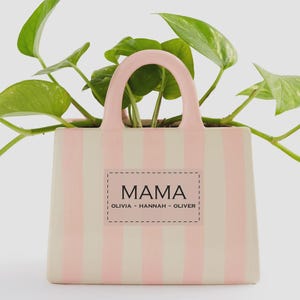 May include: A ceramic planter shaped like a tote bag, with vertical pink stripes on a cream background. The planter has a pink handle and the word "MAMA" is displayed on a rectangular label, along with the names Olivia, Hannah, and Oliver.
