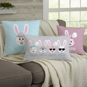 May include: Four decorative Easter-themed pillows on a gray sofa. The pillows feature cartoon bunny faces with accessories like hats and sunglasses. The pillows are light blue, pink, and gray, with names printed below the bunny faces. The pillows are decorated with white polka dots.