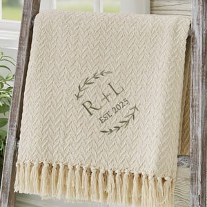 May include: Cream-colored throw blanket with a textured chevron pattern and fringe trim. The blanket features embroidered initials "R + L" within a wreath design, along with "EST 2025". The blanket is draped over a wooden ladder.