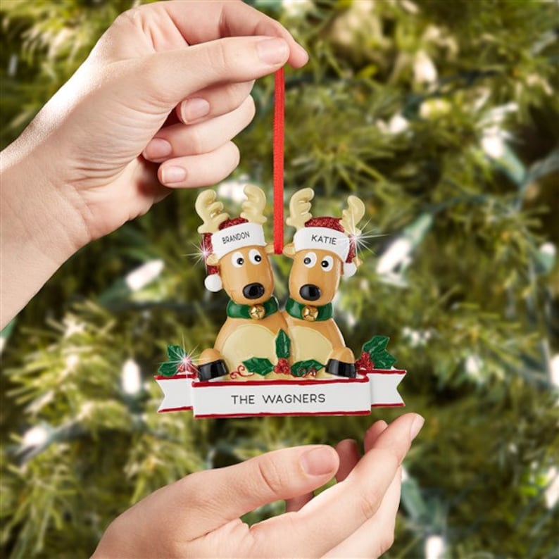 K&ouml;nnte beinhalten: Weihnachtsschmuck mit zwei Rentieren, die festliche H&uuml;te und Glocken tragen. Auf den H&uuml;ten stehen die Namen "Brandon" und "Katie". Ein Banner darunter zeigt "THE WAGNERS". Der Schmuck wird von einem roten Band gehalten.