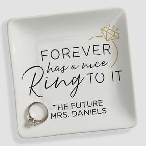 May include: A square white ceramic trinket dish featuring a gold line drawing of a diamond ring and black text that reads FOREVER has a nice RING TO IT and THE FUTURE MRS. DANIELS. A silver-toned metal ring rests on the bottom left corner.