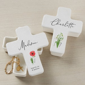 May include: Two white ceramic cross-shaped boxes with floral designs. The box on the left has the name "Madison" and a red poppy with the date "8.27.14". The box on the right has the name "Charlotte" and white snowdrops with the date "1.12.08".