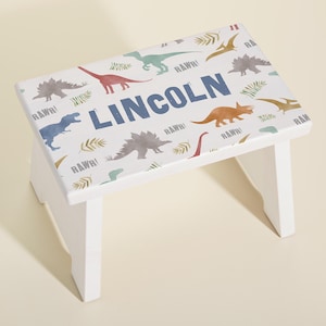 May include: White wooden step stool with a dinosaur theme. The stool is decorated with colorful dinosaurs, leaves, and the word "RAWR!" The name "LINCOLN" is printed in blue on the top of the stool.