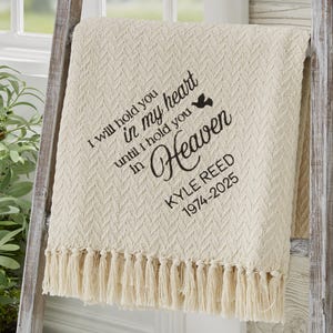 May include: Cream-colored throw blanket with a textured pattern and fringe. The blanket has the text "I will hold you in my heart until I hold you in Heaven" and the name "KYLE REED 1974-2025" in black script.