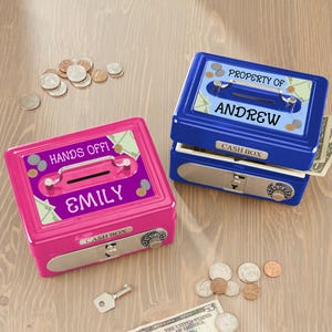 May include: Two personalized cash boxes, one pink and one blue, sit on a wooden surface. The pink box reads "HANDS OFF!" and "EMILY." The blue box says "PROPERTY OF ANDREW." Both have a slot for money, a combination lock, and a handle.