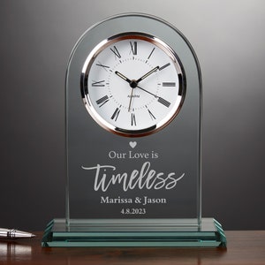 May include: A glass clock with a white face and black Roman numeral hour markers. The clock has a silver trim and a heart symbol above the inscription "Our Love is Timeless Marissa & Jason 4.8.2023".