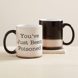 May include: Two ceramic mugs with black handles. One mug is beige with the text "You've Just Been Poisoned" in black. The other mug is black and beige with the word "Bye..." in black. Both mugs are on a light-colored surface.