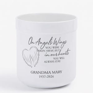 May include: White ceramic memorial candle holder with the text "On Angel's Wings" and a wing graphic. The text continues with "You were taken away, but in our hearts you will always stay." The name "Grandma Mary" and dates 1937-2026 are also printed.