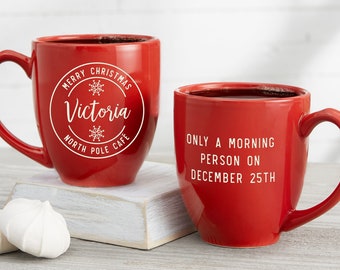 May include: Two red ceramic mugs with black liquid inside. One mug has the text "MERRY CHRISTMAS Victoria NORTH POLE CAFE" in white. The other mug has the text "ONLY A MORNING PERSON ON DECEMBER 25TH" in white.