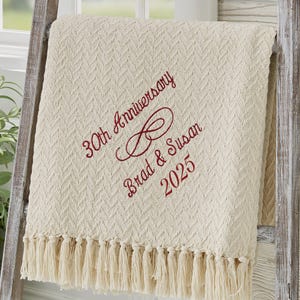 May include: Cream-colored throw blanket with a textured pattern and fringe. Embroidered in burgundy are the words "30th Anniversary", "Brad & Susan", and the year "2025". The blanket is draped over a wooden ladder.