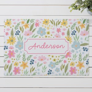 May include: A rectangular mat featuring a white background with a colorful floral pattern of yellow, pink, and light blue flowers, green leaves, and dark blue berries. A central pink-bordered rectangular frame contains the name Anderson.