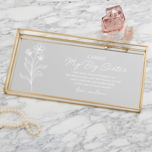 May include: A gold-rimmed glass tray with a mirrored surface. The tray has a white floral design and a personalized message that reads "CARRIE My Big Sister You have always brought out the best in me, saw the beauty in me, and loved me unconditionally. I love you so very much! love, melanie"