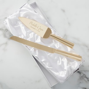May include: A gold-colored cake knife and server set, presented in a white box with a satin lining. The server is engraved with "Michelle & Jason" and "December 4, 2024". The set is designed for weddings or special occasions.