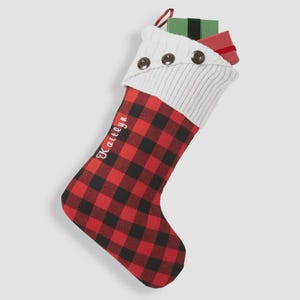 May include: A Christmas stocking with a red and black buffalo plaid pattern. The top is white knit with three brown buttons. The name "Kaitlyn" is written in cursive. Two wrapped gifts are inside.