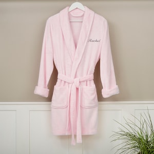 May include: A light pink terry cloth robe with a shawl collar, long sleeves, and a matching belt. The robe has two front pockets and the name "Rachel" embroidered on the chest. It is hanging on a white hanger.