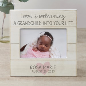 May include: White wooden picture frame with the text "Love is welcoming a grandchild into your life". The frame has a photo of a baby girl with a pink headband and a pink blanket. The text "Rosa Marie August 20, 2023" is below the photo with a pink heart.