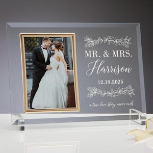 May include: A clear glass photo frame with a gold-colored border. The frame holds a wedding photo of a couple. The text "MR. & MRS. Harrison 12.19.2025 a true love story never ends" is printed on the frame.