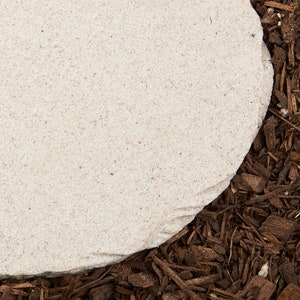 May include: A light beige, rounded stone paver with a rough texture, set against a backdrop of dark brown wood chips. The stone's edge is visible, showing its irregular shape and natural color variations. Ideal for garden or landscaping projects.