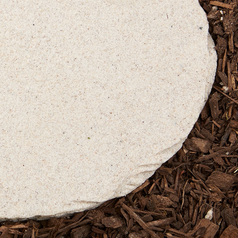 May include: A light beige, rounded stone paver with a rough texture, set against a backdrop of dark brown wood chips. The stone's edge is visible, showing its irregular shape and natural color variations. Ideal for garden or landscaping projects.