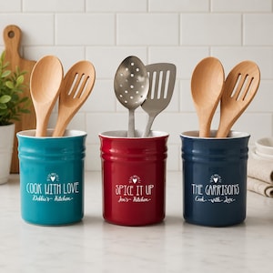 May include: Three ceramic kitchen utensil holders in teal, red, and navy blue. Each holder has a white interior and features a personalized message in white script. Wooden spoons and metal spatulas are inside the holders.