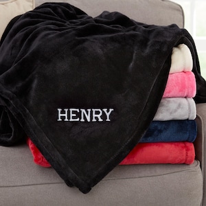 May include: A black plush blanket with the name "HENRY" embroidered in white. The blanket is draped over a stack of folded blankets in various colors, including white, pink, gray, navy blue, and red. The blankets are on a gray sofa.