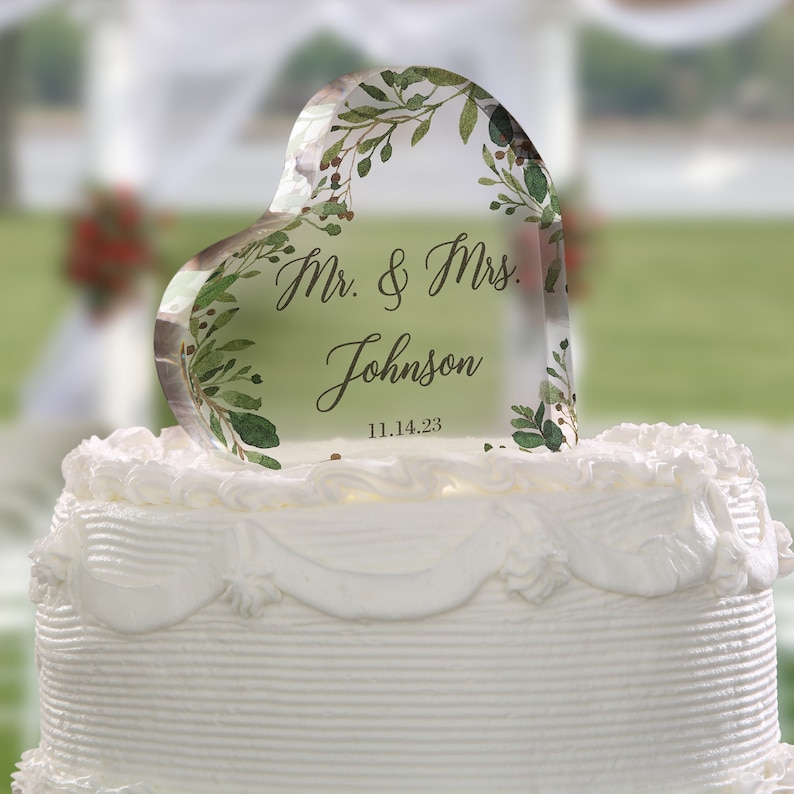 May include: A clear heart-shaped cake topper with a watercolor design of green leaves and berries. The topper is personalized with the text "Mr. & Mrs. Johnson" and the date "11.14.23".