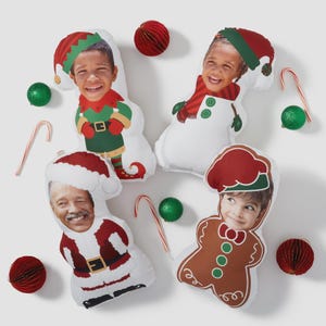 May include: Four custom-shaped throw pillows featuring photographic faces of individuals printed onto bodies dressed as a Christmas elf, a snowman, Santa Claus, and a gingerbread person, surrounded by small candy canes and red and green ornaments.