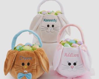 May include: Three plush bunny-shaped Easter baskets with handles, filled with colorful plastic eggs and green paper grass. The baskets are personalized with embroidered names: Kennedy in teal, Owen in blue, and Addison in pink, featuring bunny faces.