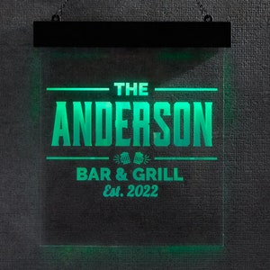 May include: A rectangular, illuminated sign with the text "THE ANDERSON BAR & GRILL Est. 2022" in bright green. The sign is suspended by a black metal frame and chain. The background is a dark gray textured wall.