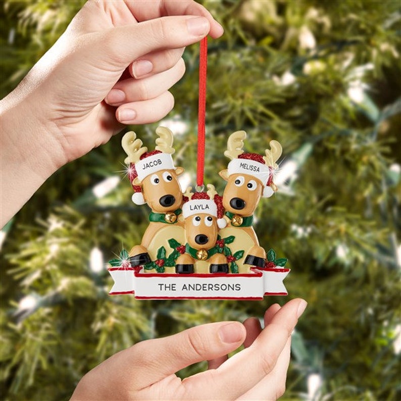 K&ouml;nnte beinhalten: Weihnachtsschmuck mit drei Rentieren, die H&uuml;te mit den Namen "Jacob", "Layla" und "Melissa" tragen. Das Ornament hat ein Banner mit der Aufschrift "THE ANDERSONS". Die Rentiere tragen gr&uuml;ne Kragen und rot-wei&szlig;e H&uuml;te.