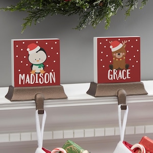 May include: Two Christmas stocking holders with red backgrounds and white polka dots. One features a penguin with a Santa hat and the name "Madison". The other has a reindeer with a Santa hat and the name "Grace".