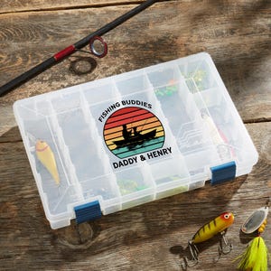 May include: A clear plastic fishing tackle box with multiple compartments, featuring a colorful sunset graphic with the text "Fishing Buddies" and "Daddy & Henry." Fishing lures and a fishing rod are also visible.