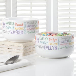 May include: Two white ceramic bowls with colorful text and bunny graphics. One bowl says "MAVERICK" in various colors, the other says "EVELYN". The bowl on the right is filled with colorful cereal.