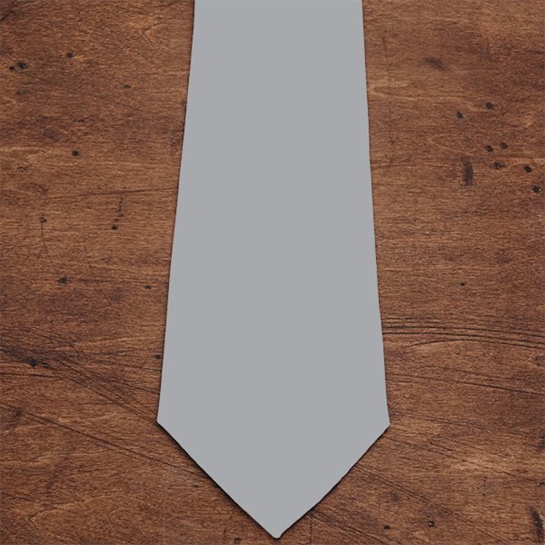 May include: A plain grey necktie, shown against a wooden background. The tie is a light shade of grey, with a traditional pointed tip. The backdrop has a warm, wooden texture.