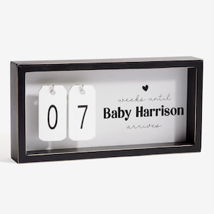 May include: A black framed wooden sign with the text "weeks until Baby Harrison arrives." Two white tags display the numbers "0" and "7". A small black heart is above the text. The sign is a countdown to a baby's arrival.